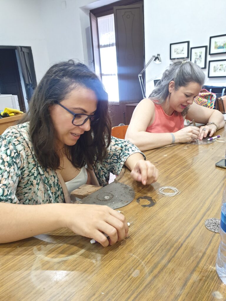 Dos participantes trabajando el hilo de plata en el taller; sobre la mesa, una pieza de filigrana terminada
